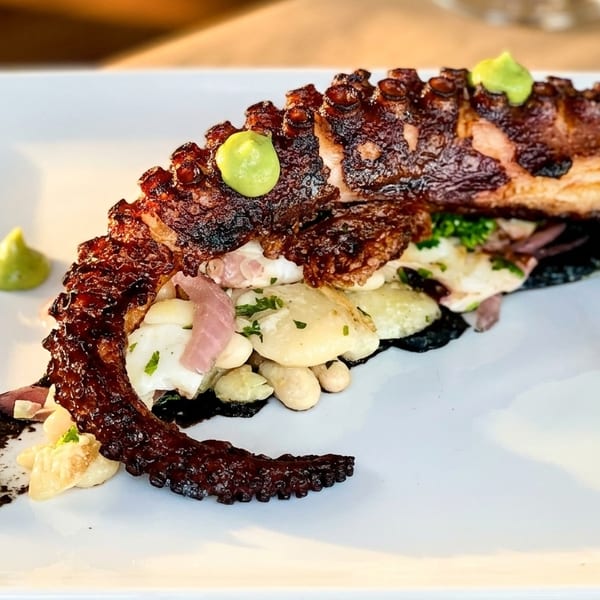 Grilled octopus with white beans, red onions, and green sauce on a white rectangular plate.
