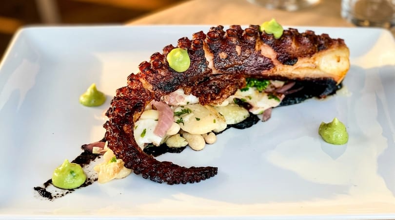 Grilled octopus with white beans, red onions, and green sauce on a white rectangular plate.