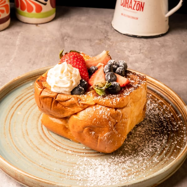 Golden French toast with whipped cream, strawberries, blueberries, and powdered sugar on a textured plate.