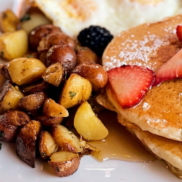 A breakfast plate with pancakes, strawberries, blackberries, roasted potatoes, and fried eggs.