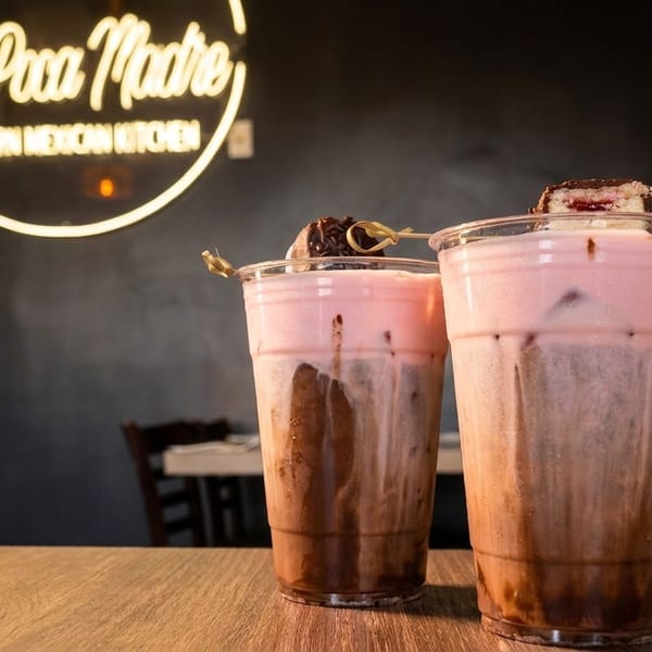 Two layered chocolate and pink drinks with toppings on a wooden table, under a neon sign.
