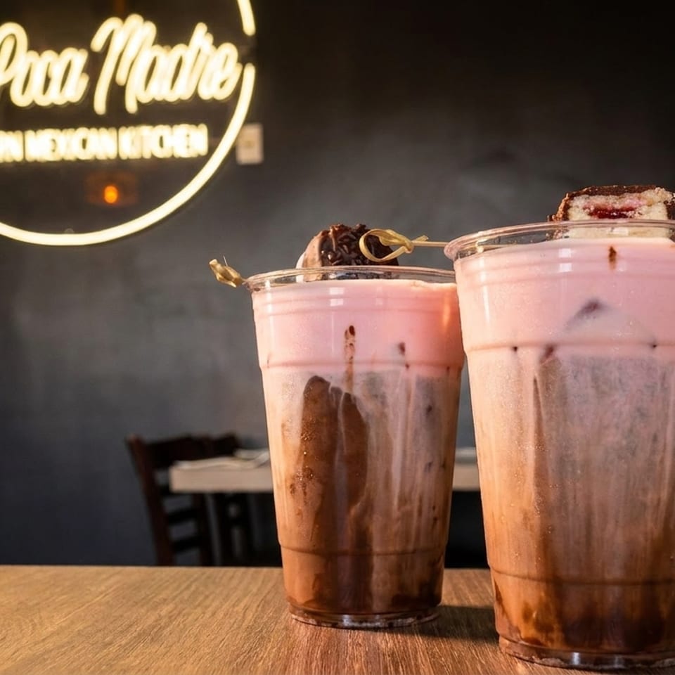 Two layered chocolate and pink drinks with toppings on a wooden table, under a neon sign.