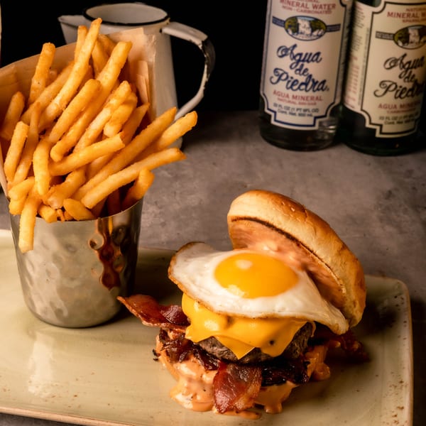 Juicy burger topped with a fried egg, bacon, and cheese, alongside crispy fries and mineral water.