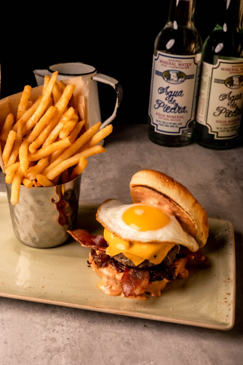 Juicy burger topped with a fried egg, bacon, and cheese, alongside crispy fries and mineral water.