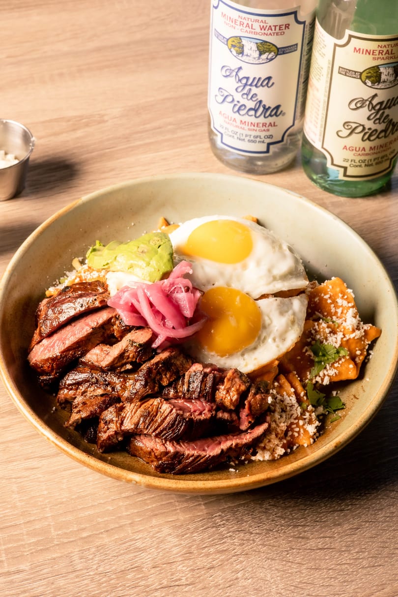 Bowl of chilaquiles with sliced steak, two fried eggs, avocado, pickled onions, and mineral water bottles.